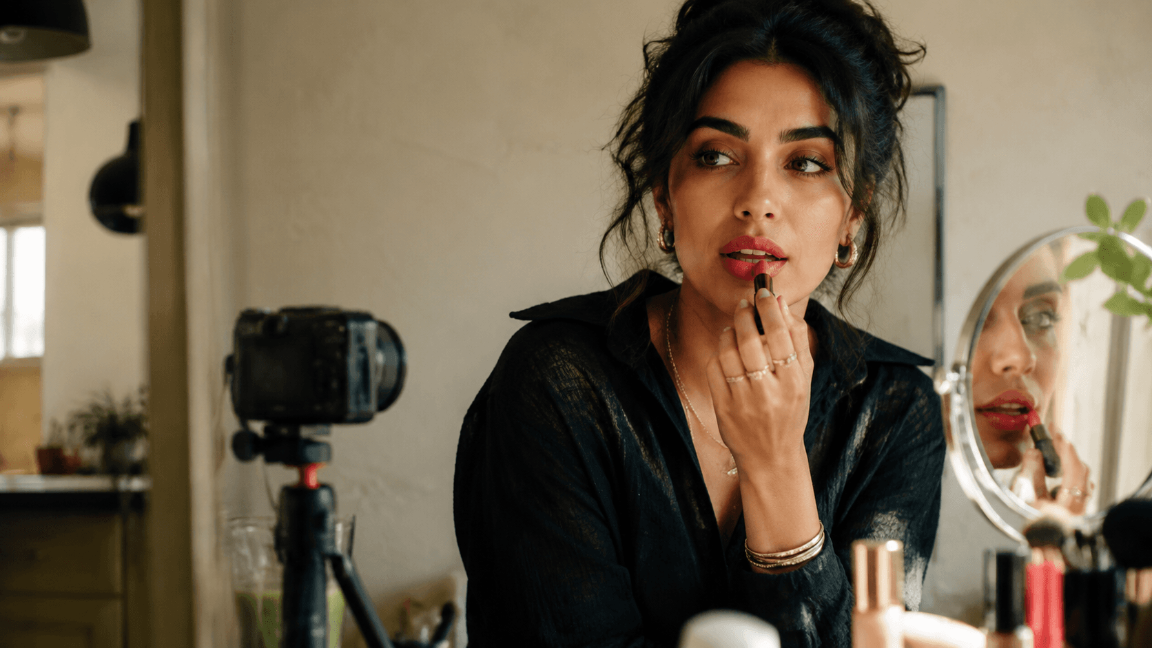 Temporary editorial creator portrait: a woman applying lipstick at a sunlit vanity beside a tripod-mounted camera